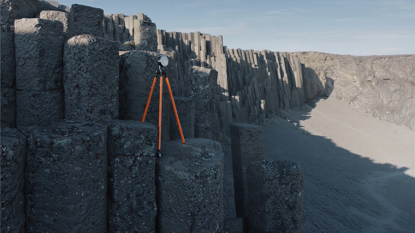 Tall hexagonal basalt columns in low morning sun, an orange survey tripod with a round prism standing on a column top, the columned valley wall receding along a pale gravel floor