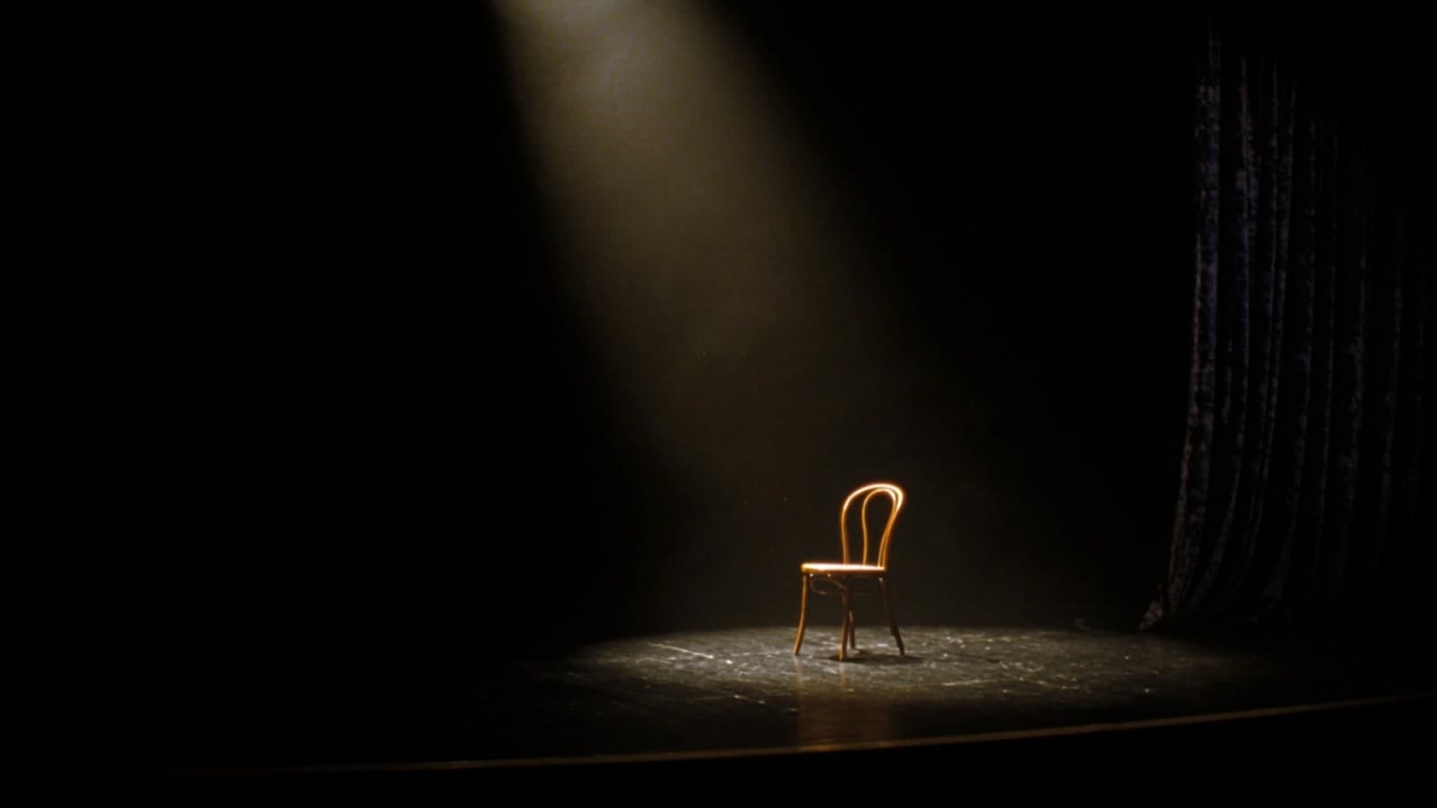 A bentwood chair under a single hard spotlight on an empty black stage, haze in the beam and a dark velvet curtain at the right