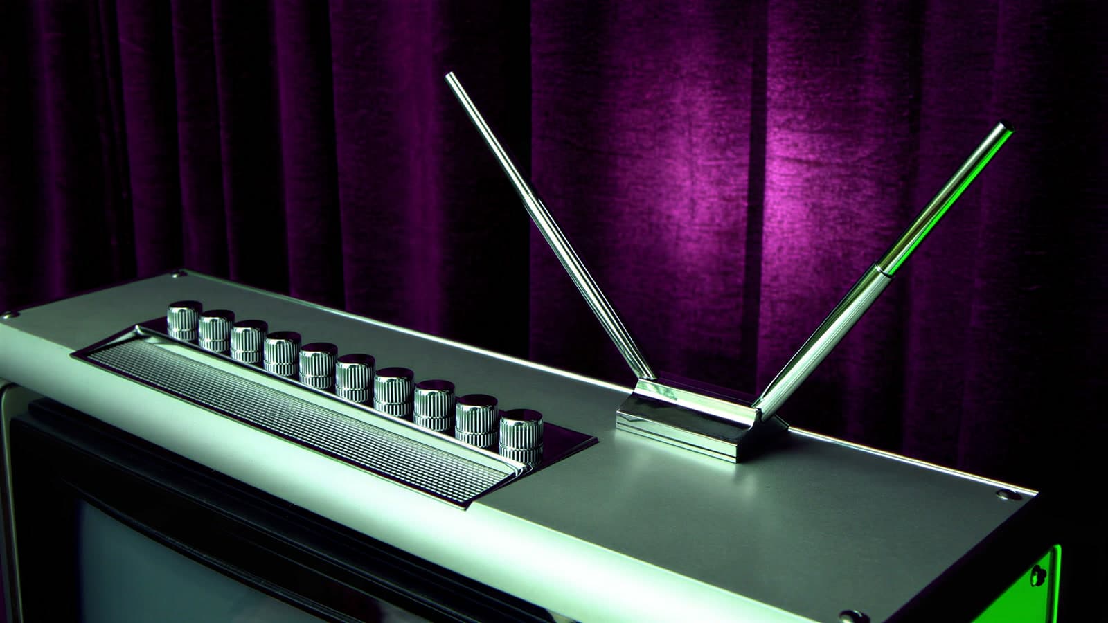 The top of a brushed aluminium television with a row of chrome tuning knobs and a chrome rabbit-ear antenna, a green rim of light on its edge, against a deep violet velvet drape